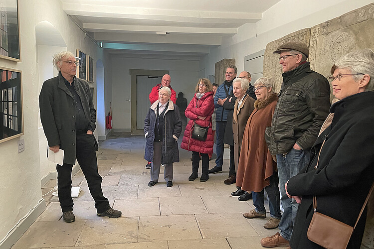 Gang in einem historischen Gebäude mit steinernen Grabplatten; Teilnehmende einer Führung hören einem Mann zu