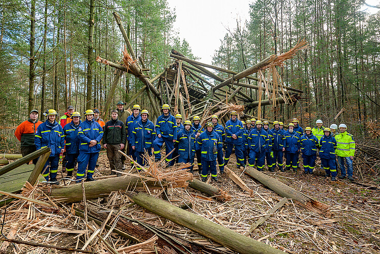 Eine Gruppe von Einsatzkräften des Technischen Hilfswerks sowie Mitarbeiter der Klosterforsten stehen gemeinsam im Wald vor dem gesprengten hölzernen Feuerwachturm.