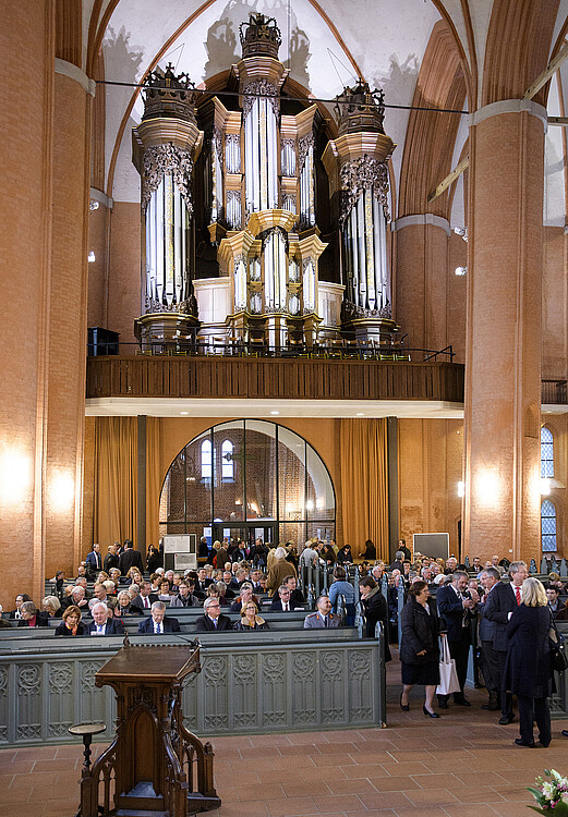 Blick in einen Kircheninnenraum mit Personen, die in den Bänken sitzen und einer großen Orgel im Hintergrund.