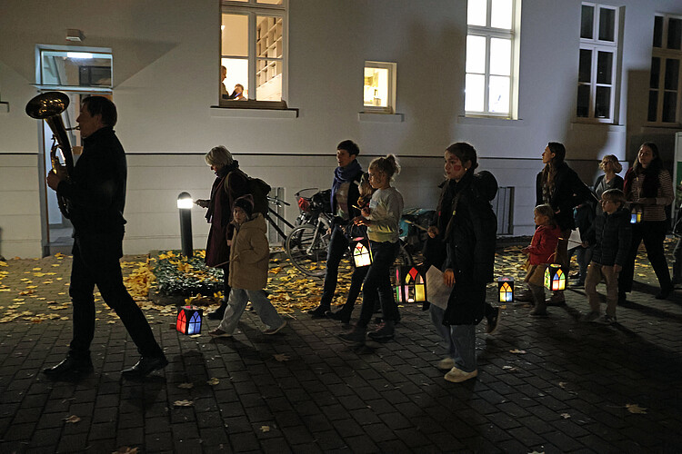 Ein Mann, der auf einem Tenorhorn spielt, führt einen Laternen-Umzug über den Hof der Klosterkammer an. Ihm folgen Erwachsene und Kinder mit Laternen aus buntem Papier.