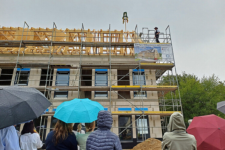 Eine Gruppe von Menschen ist von hinten - teils mit Regenschirmen - zu sehen. Sie blicken zu einem Zimmermann auf dem Gerüst vor einem Rohbau hoch.