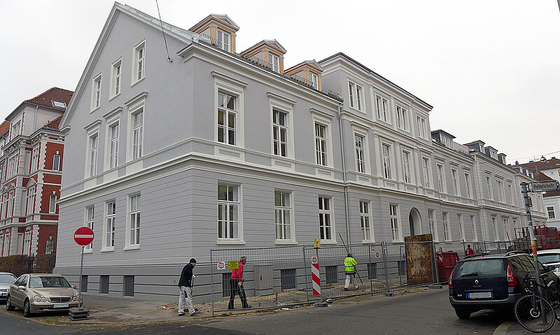 Ein rechteckiges, dreistöckiges Gebäude in Grau mit einem Ziegeldach.