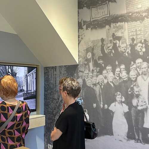 Zwei Frauen, die von hinten zu sehen sind, schauen sich eine Ausstellung mit historischem Filmmaterial und Fotos an