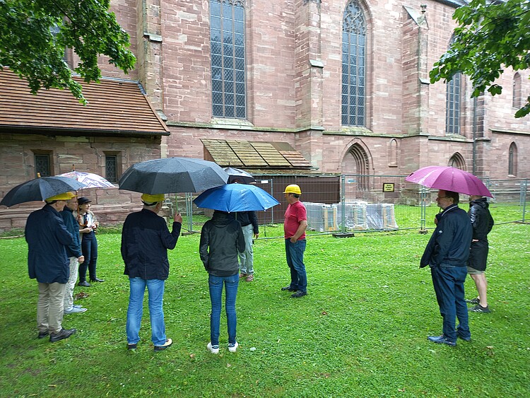 Mehrere Personen mit Bauhelmen und Regenschirmen auf einer Wiese neben einer Kirche. Kirche nur ausschnittsweise zu sehen.