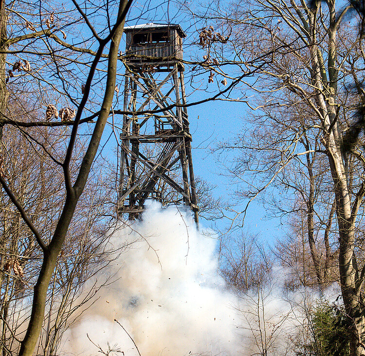 Ein Turm aus hölzernen Streben wird während der Sprengung gezeigt, im unteren Bereich ist eine Staubwolke zu sehen.