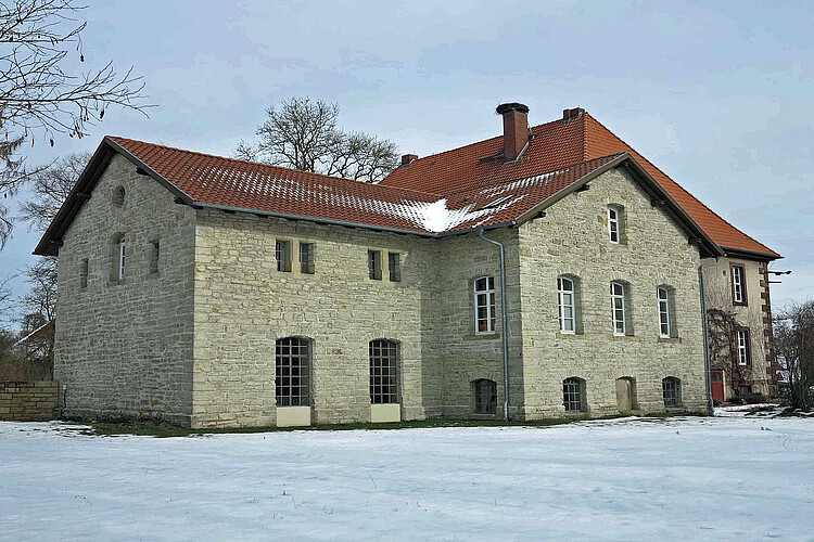 Seitenansicht eines schlichten, historischen Gebäudes mit heller Fassade und roter Dacheindeckung.