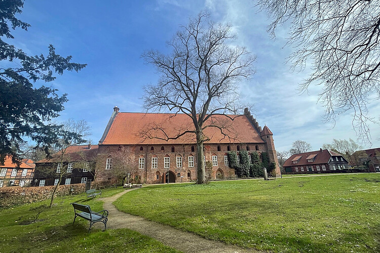 Blick auf die seitliche Backsteinfassade des Klosters Wienhausen. Davor ist ein Teil des Parks mit Wiese, Bänken und Bäumen zu sehen.